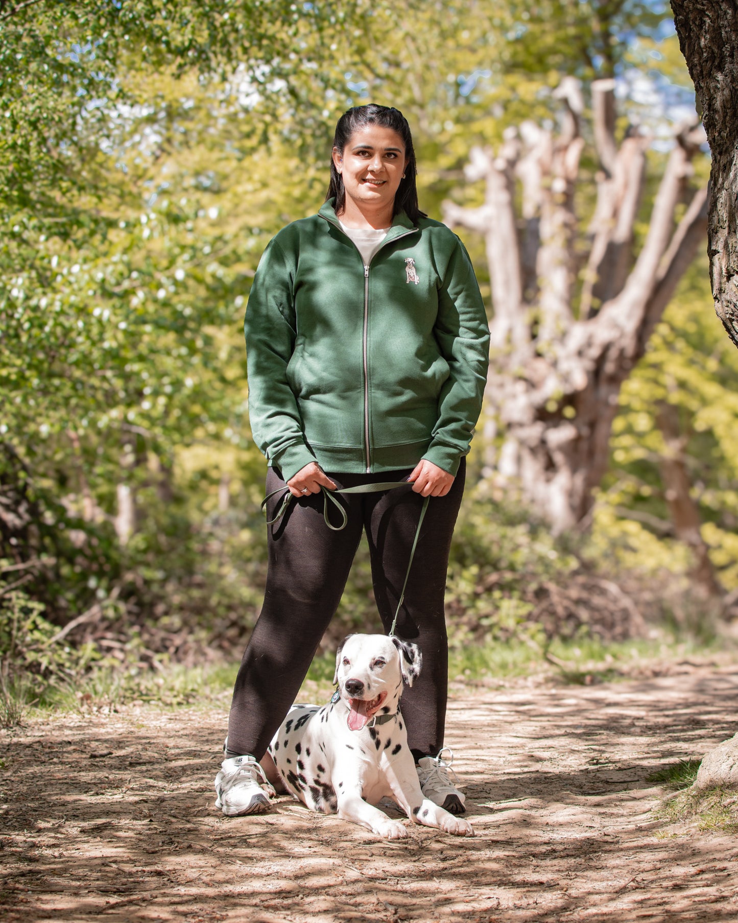 Woman in a green jacket standing with a Dalmatian dog on a path in a park.