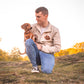 Man sitting on a grassy hill with a Miniature Dachshund, wearing a jacket with a pet portrait design.