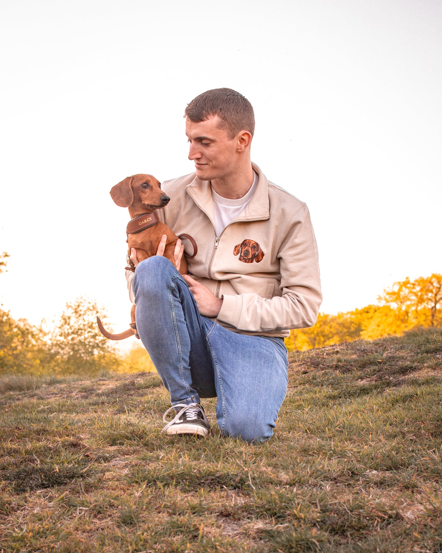 Man sitting on a grassy hill with a Miniature Dachshund, wearing a jacket with a pet portrait design.